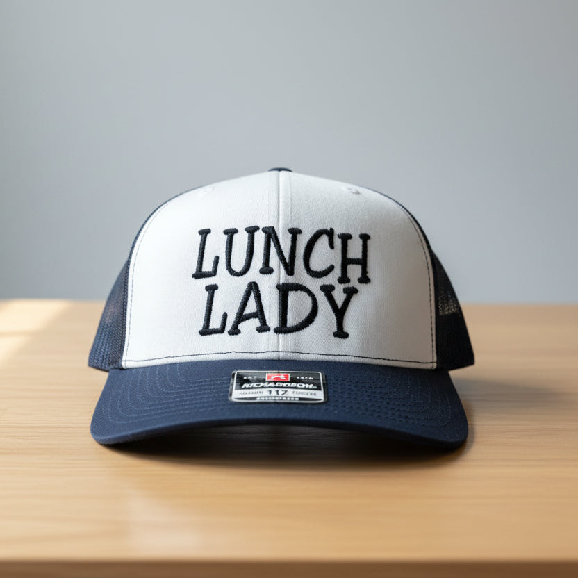 Trucker hat with 'LUNCH LADY' text on a wooden surface and gray background